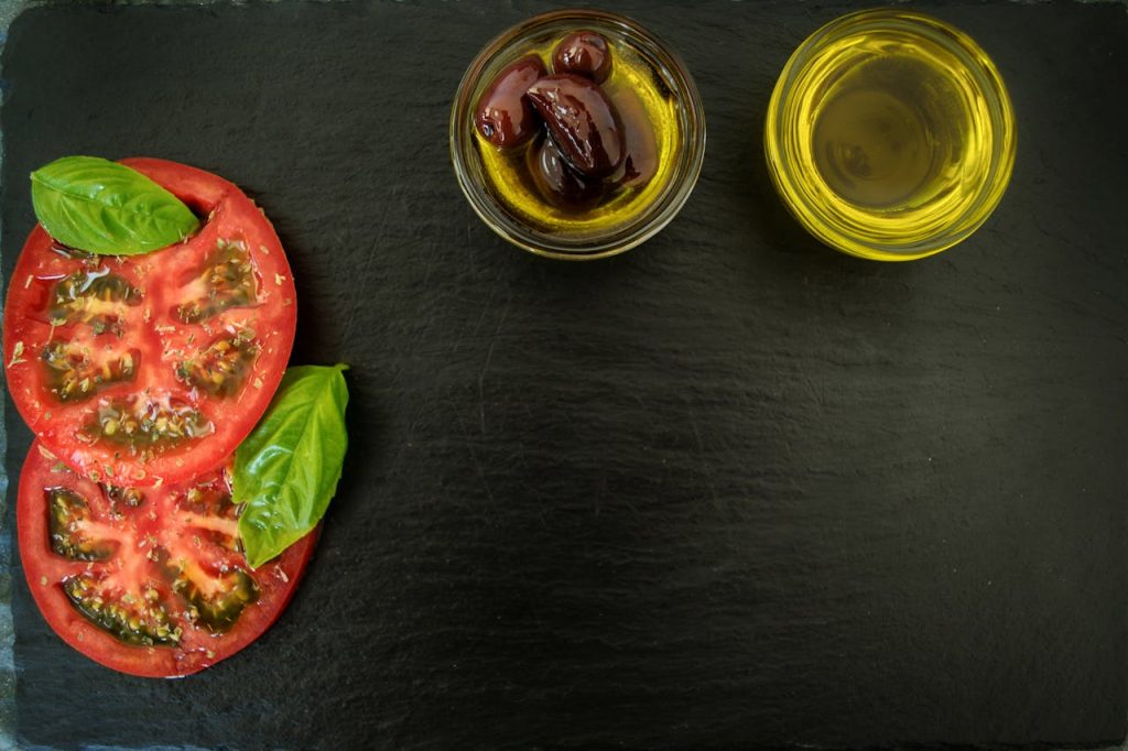 Top view of fresh basil, sliced tomatoes, olives, and olive oil on slate, perfect for Italian cuisine themes.